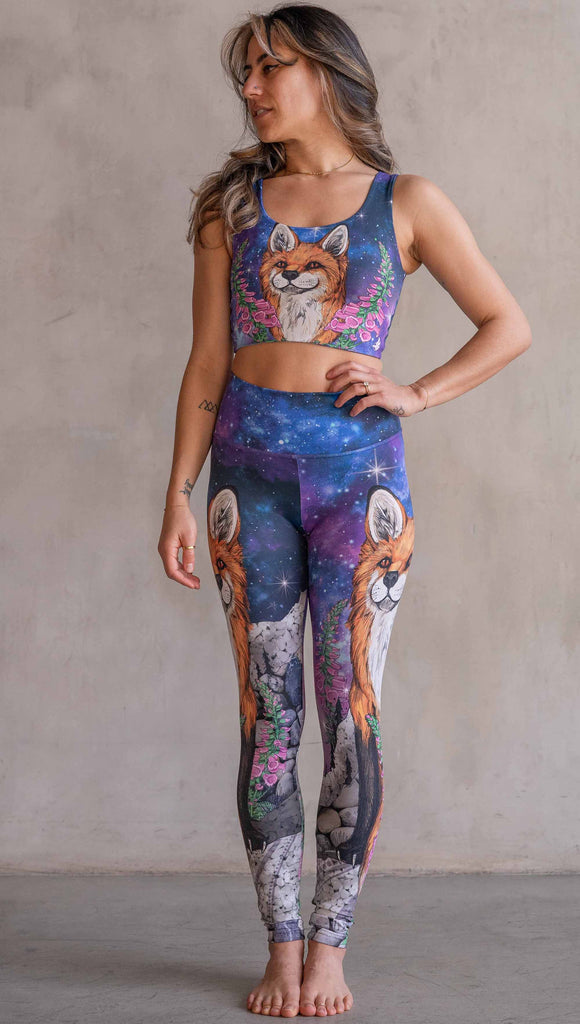 Girl wearing WERKSHOP X Save A Fox Athleisure Leggings. The artwork on the leggings features a coy red fox looking upward while perched on a rock and framed with foxglove flowers. The background for the artwork features a galactic sky with swirls of purple and blue and sparkling stars.