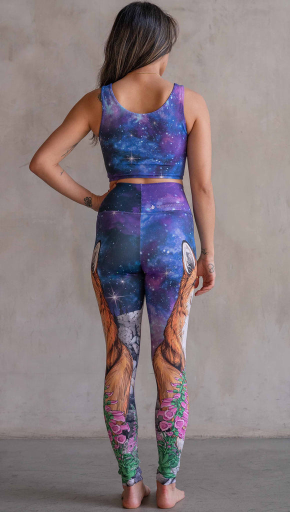 Girl wearing WERKSHOP X Save A Fox Athleisure Leggings. The artwork on the leggings features a coy red fox looking upward while perched on a rock and framed with foxglove flowers. The background for the artwork features a galactic sky with swirls of purple and blue and sparkling stars.