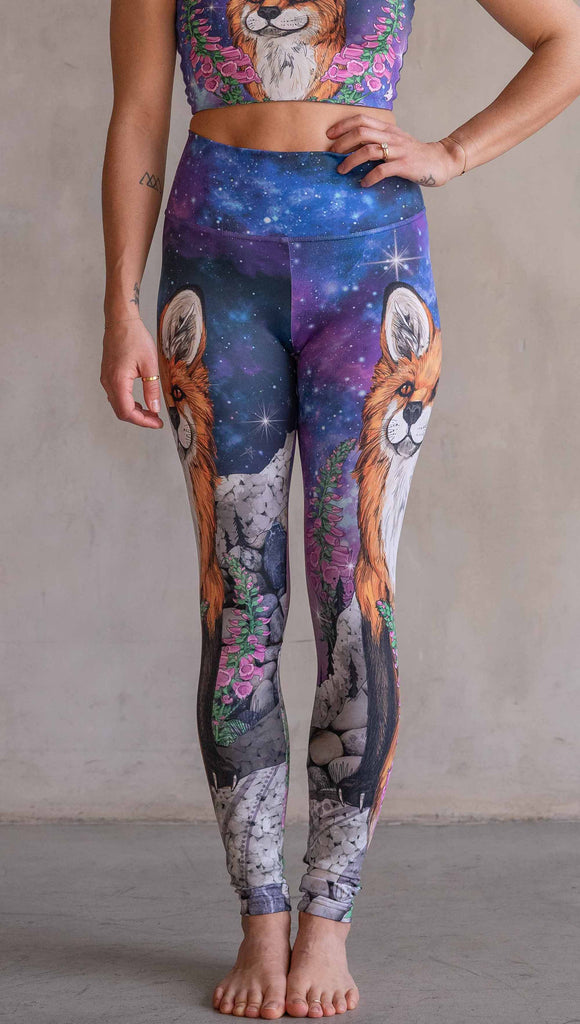 Girl wearing WERKSHOP X Save A Fox Athleisure Leggings. The artwork on the leggings features a coy red fox looking upward while perched on a rock and framed with foxglove flowers. The background for the artwork features a galactic sky with swirls of purple and blue and sparkling stars.