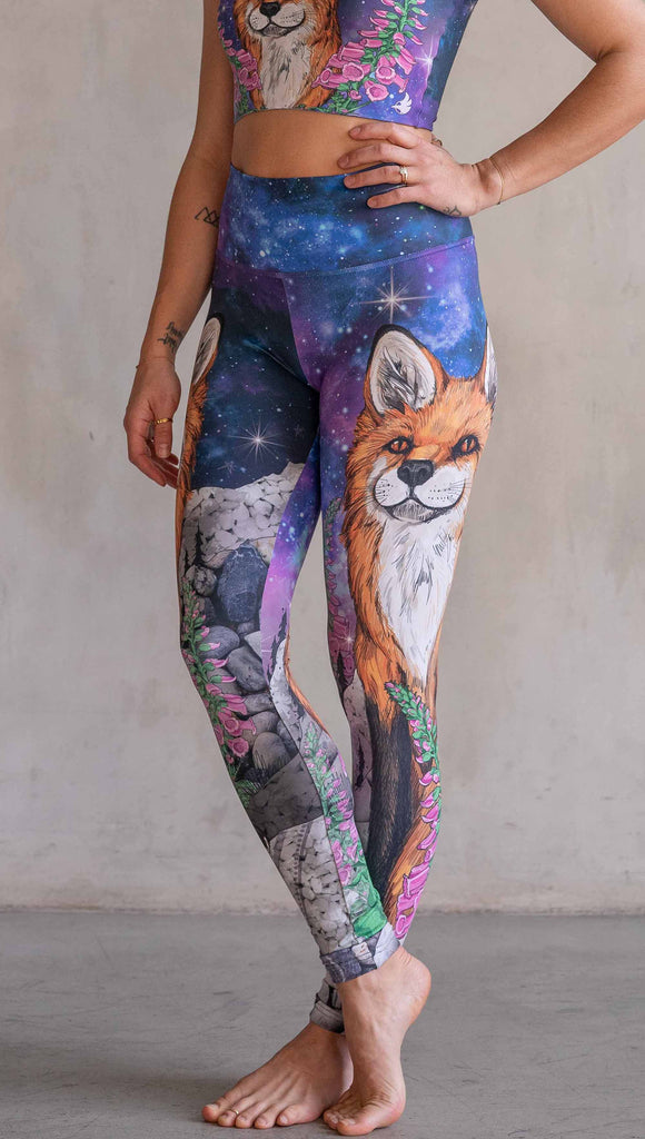 Girl wearing WERKSHOP X Save A Fox Athleisure Leggings. The artwork on the leggings features a coy red fox looking upward while perched on a rock and framed with foxglove flowers. The background for the artwork features a galactic sky with swirls of purple and blue and sparkling stars.