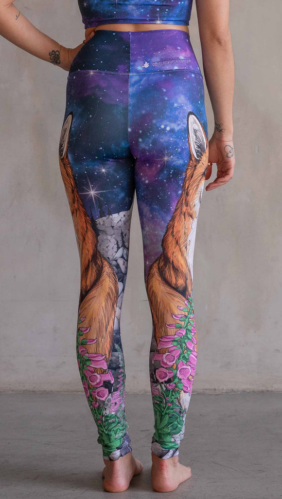 Girl wearing WERKSHOP X Save A Fox Athleisure Leggings. The artwork on the leggings features a coy red fox looking upward while perched on a rock and framed with foxglove flowers. The background for the artwork features a galactic sky with swirls of purple and blue and sparkling stars.