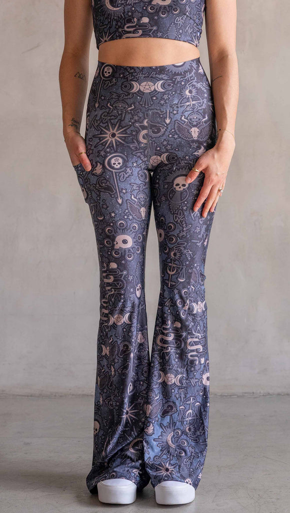 Model wearing WERKSHOP Tarot Bell Bottoms. They are high waist and feature pockets on both legs. The blue/tarot artwork has skulls, snakes, moons and the names of multiple popular tarot cards like "Strength, Lovers, Death and The Hanged Man”. Our model is 5’6” and this images shows her wearing the bells barefoot. The hem of the pants are hitting the floor.