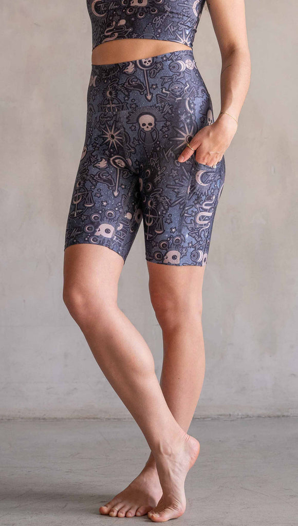 Model wearing WERKSHOP Tarot Bicycle Length Shorts. They are high waist and feature pockets on both legs. The blue/tarot artwork has skulls, snakes, moons and the names of multiple popular tarot cards like "Strength, Lovers, Death and The Hanged Man”. The length of the shorts hit in the mid thigh, above the knee.