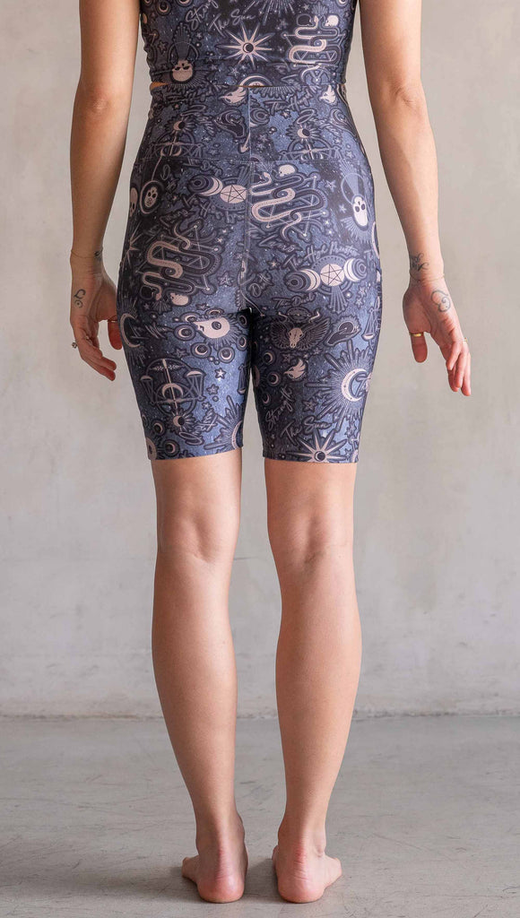 Model wearing WERKSHOP Tarot Bicycle Length Shorts. They are high waist and feature pockets on both legs. The blue/tarot artwork has skulls, snakes, moons and the names of multiple popular tarot cards like "Strength, Lovers, Death and The Hanged Man”. The length of the shorts hit in the mid thigh, above the knee.