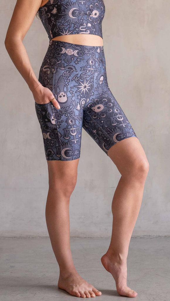 Model wearing WERKSHOP Tarot Bicycle Length Shorts. They are high waist and feature pockets on both legs. The blue/tarot artwork has skulls, snakes, moons and the names of multiple popular tarot cards like "Strength, Lovers, Death and The Hanged Man”. The length of the shorts hit in the mid thigh, above the knee.