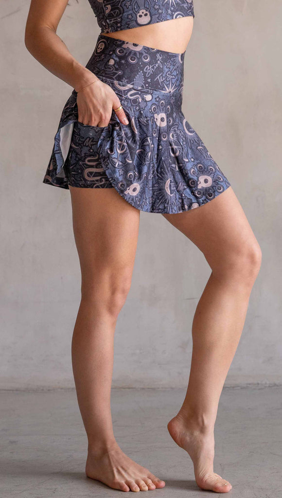 Model wearing WERKSHOP Zodiac Tennis Skirt with built-in shorts. They are high waist and feature pockets on both legs under the flirty skirt. The blue/tarot artwork has skulls, snakes, moons and the names of multiple popular tarot cards like "Strength, Lovers, Death and The Hanged Man"