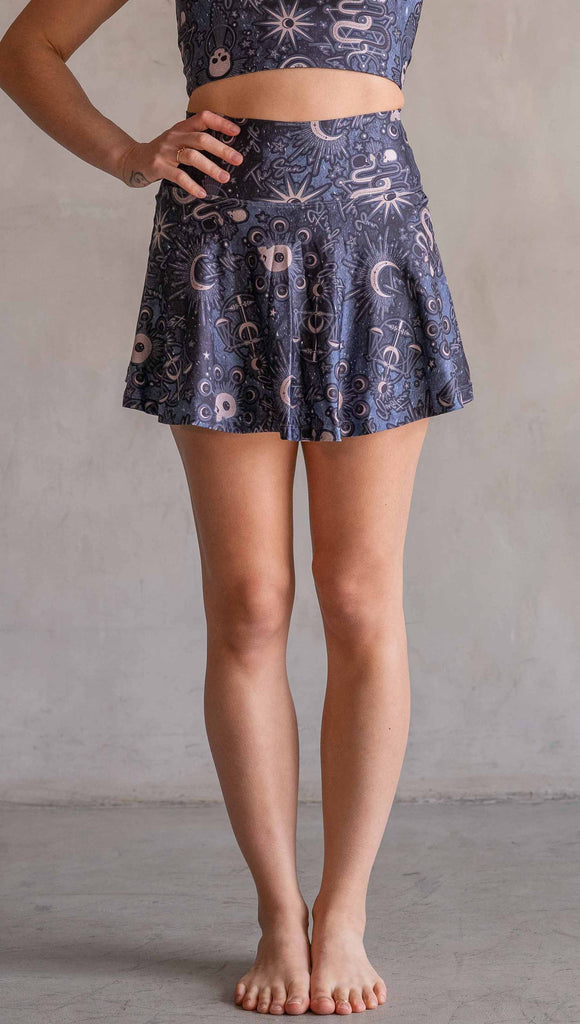 Model wearing WERKSHOP Zodiac Tennis Skirt with built-in shorts. They are high waist and feature pockets on both legs under the flirty skirt. The blue/tarot artwork has skulls, snakes, moons and the names of multiple popular tarot cards like "Strength, Lovers, Death and The Hanged Man"