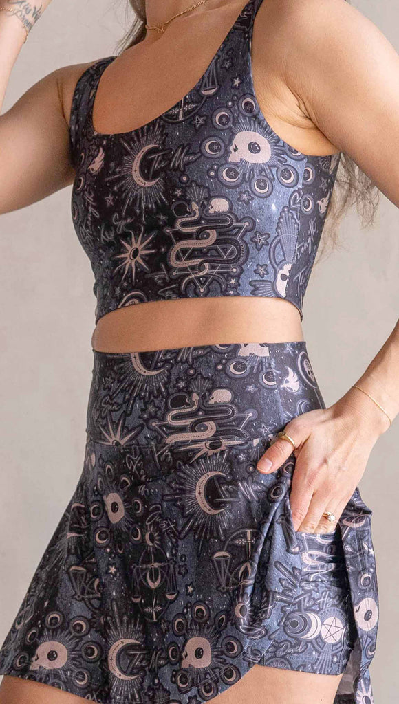 Model wearing WERKSHOP Zodiac Tennis Skirt with built-in shorts. They are high waist and feature pockets on both legs under the flirty skirt. The blue/tarot artwork has skulls, snakes, moons and the names of multiple popular tarot cards like "Strength, Lovers, Death and The Hanged Man"