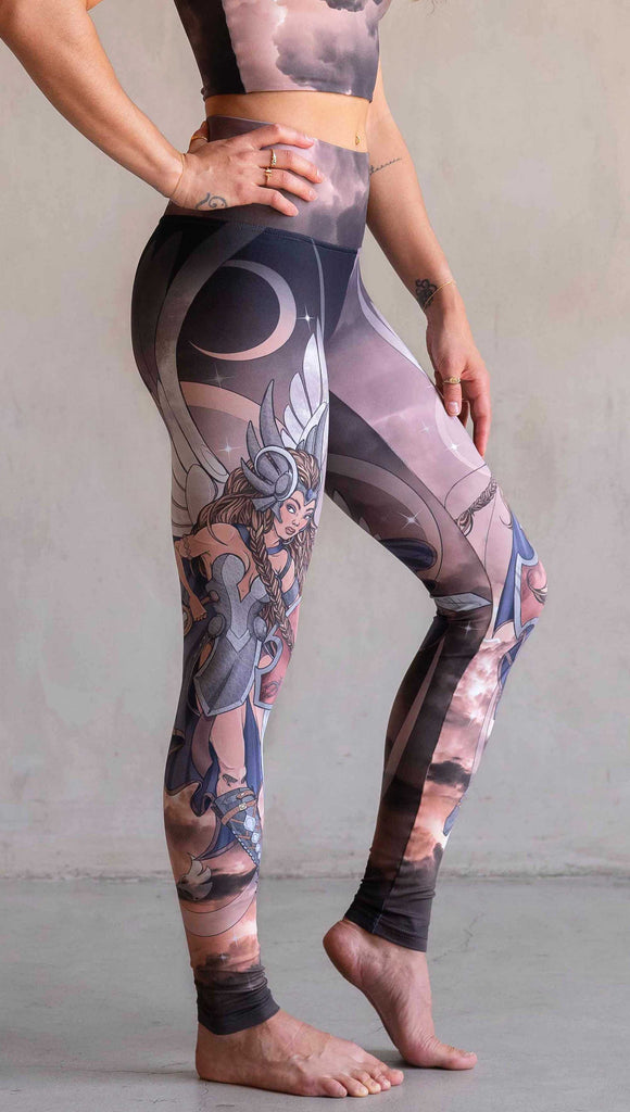 Side view of model wearing WERKSHOP Valkyrie Athleisure Leggings. featuring original artwork by Chriztina Marie. featuring a Valkyrie warrior from Norse Mythology flying through a dark sky holding a shield and swords. Her hair is in braids and she is wearing armor and a viking helmet. The colors are warm and the background also features a crescent moon and stars.