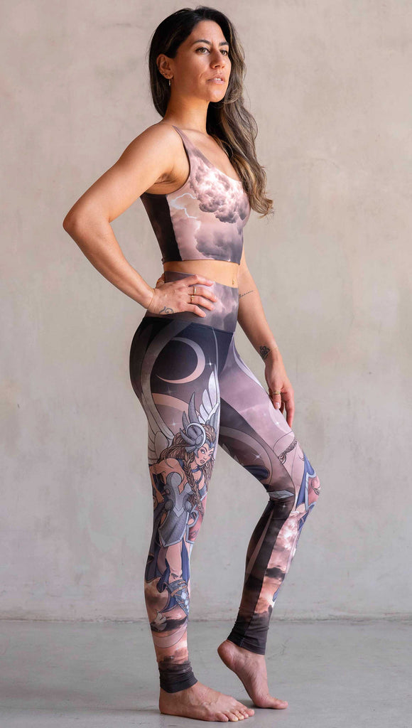 Full body side view of model wearing WERKSHOP Valkyrie Athleisure Leggings. featuring original artwork by Chriztina Marie. featuring a Valkyrie warrior from Norse Mythology flying through a dark sky holding a shield and swords. Her hair is in braids and she is wearing armor and a viking helmet. The colors are warm and the background also features a crescent moon and stars.