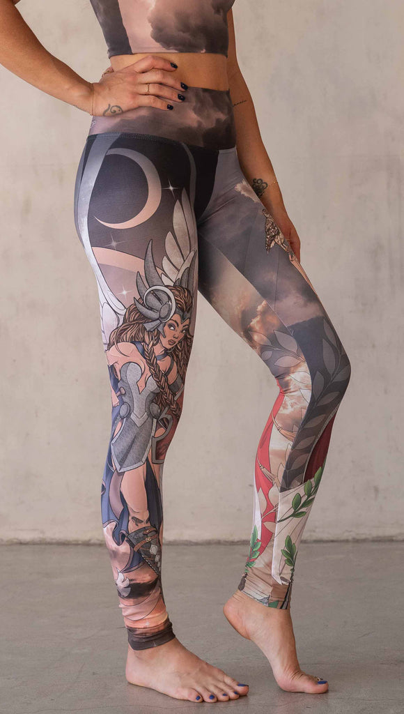 Model wearing WERKSHOP Valkyrie + Athena Mashup leggings. The leggings are printed with original artwork by Chriztina Marie. One leg features Athena, the goddess of war standing on a cliff’s edge. She is holding a spear with one hand and her owl with the other. The opposite leg features a Valkyrie warrior from Norse Mythology flying through a dark sky holding a shield and swords. Her hair is in braids and she is wearing armor and a viking helmet.