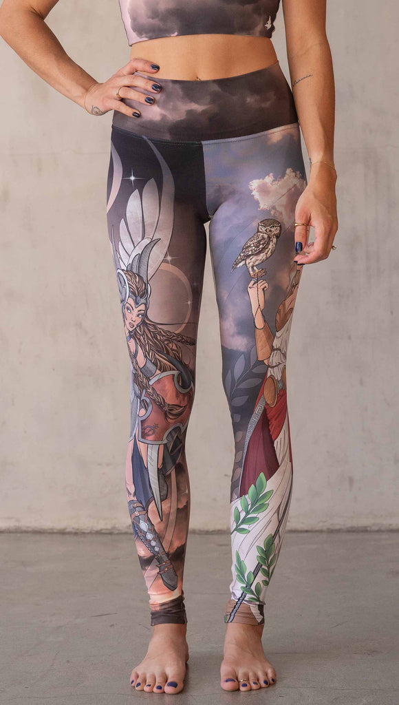 Model wearing WERKSHOP Valkyrie + Athena Mashup leggings. The leggings are printed with original artwork by Chriztina Marie. One leg features Athena, the goddess of war standing on a cliff’s edge. She is holding a spear with one hand and her owl with the other. The opposite leg features a Valkyrie warrior from Norse Mythology flying through a dark sky holding a shield and swords. Her hair is in braids and she is wearing armor and a viking helmet.