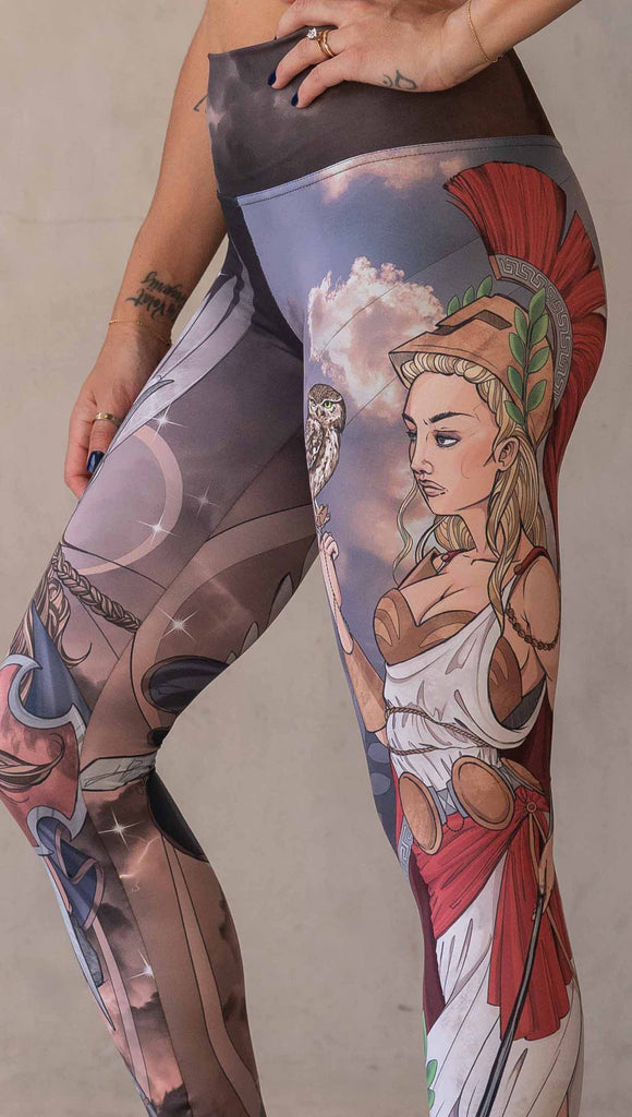 Model wearing WERKSHOP Valkyrie + Athena Mashup leggings. The leggings are printed with original artwork by Chriztina Marie. One leg features Athena, the goddess of war standing on a cliff’s edge. She is holding a spear with one hand and her owl with the other. The opposite leg features a Valkyrie warrior from Norse Mythology flying through a dark sky holding a shield and swords. Her hair is in braids and she is wearing armor and a viking helmet.