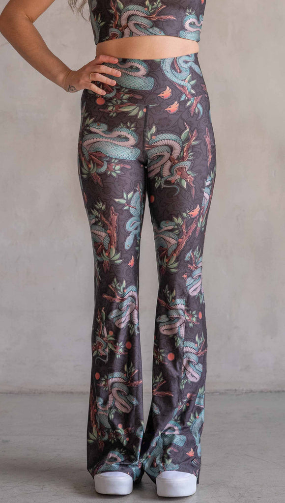 Model wearing WERKSHOP Pit Viper Featherlight Bells. The artwork on the leggings have clusters of teal blue pit viper snakes intertwined on tree branches over a taupe/brown background. The featherlight bells have pockets.