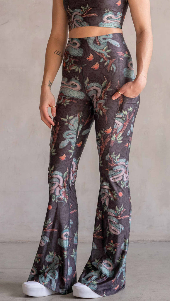 Model wearing WERKSHOP Pit Viper Featherlight Bells. The artwork on the leggings have clusters of teal blue pit viper snakes intertwined on tree branches over a taupe/brown background. The featherlight bells have pockets.