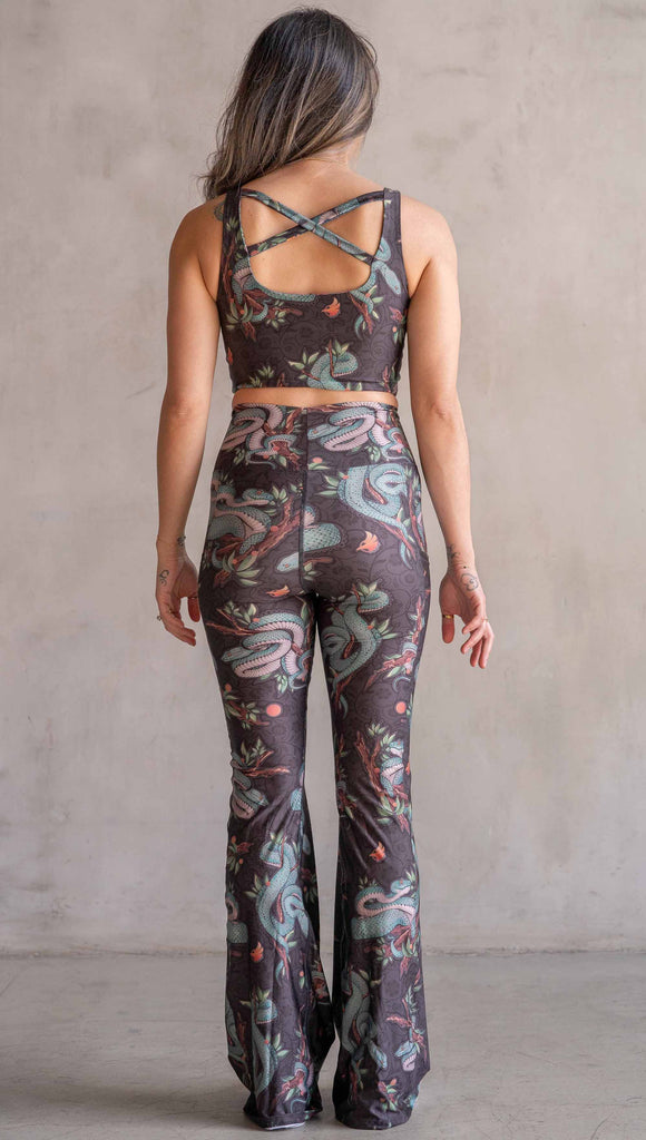 Model wearing WERKSHOP Pit Viper Featherlight Bells. The artwork on the leggings have clusters of teal blue pit viper snakes intertwined on tree branches over a taupe/brown background. The featherlight bells have pockets.