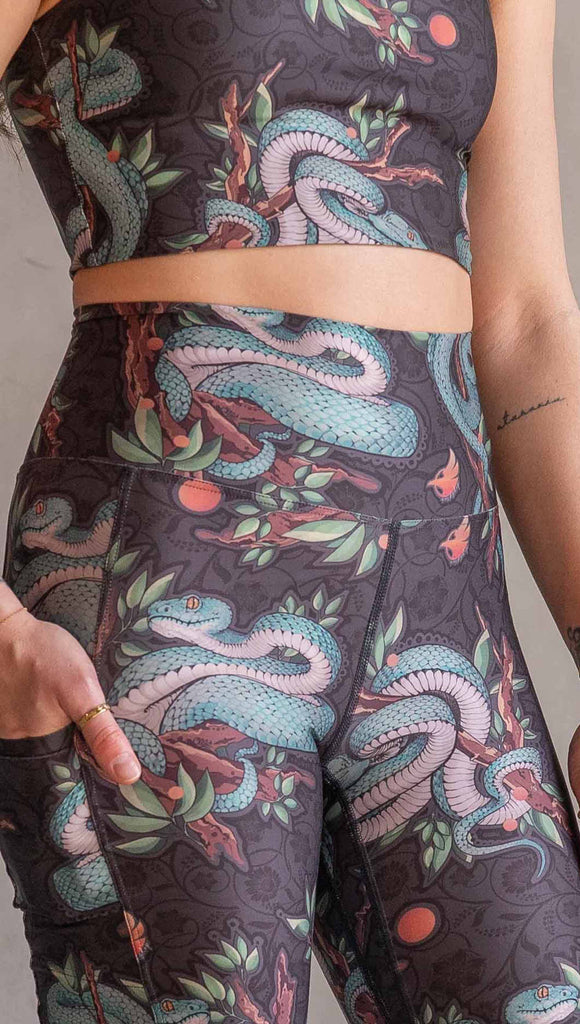 Model wearing WERKSHOP Pit Viper Featherlight Bells. The artwork on the leggings have clusters of teal blue pit viper snakes intertwined on tree branches over a taupe/brown background. The featherlight bells have pockets.