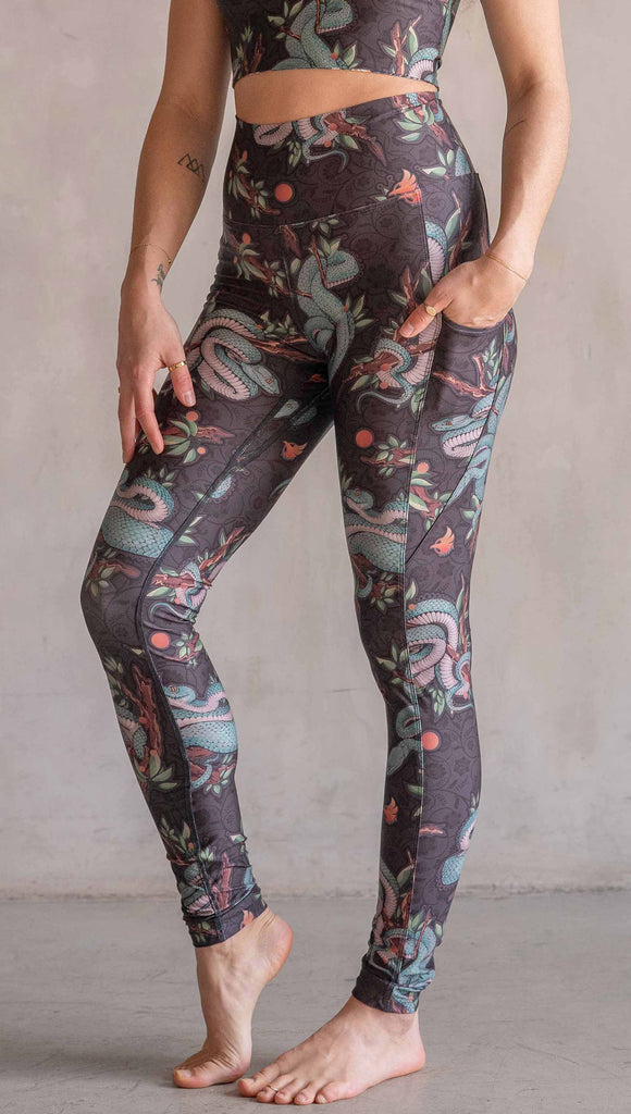 Model wearing WERKSHOP Pit Viper Featherlight Leggings. The artwork on the leggings have clusters of teal blue pit viper snakes intertwined on tree branches over a taupe/brown background. The featherlight leggings have pockets.