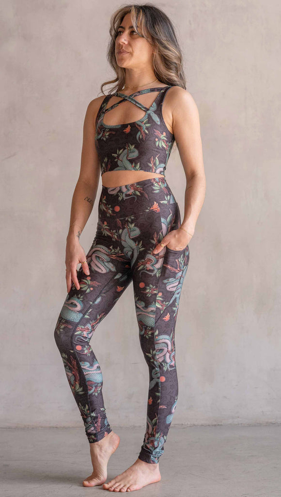 Model wearing WERKSHOP Pit Viper Featherlight Leggings. The artwork on the leggings have clusters of teal blue pit viper snakes intertwined on tree branches over a taupe/brown background. The featherlight leggings have pockets.