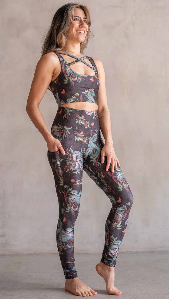 Model wearing WERKSHOP Pit Viper Featherlight Leggings. The artwork on the leggings have clusters of teal blue pit viper snakes intertwined on tree branches over a taupe/brown background. The featherlight leggings have pockets.