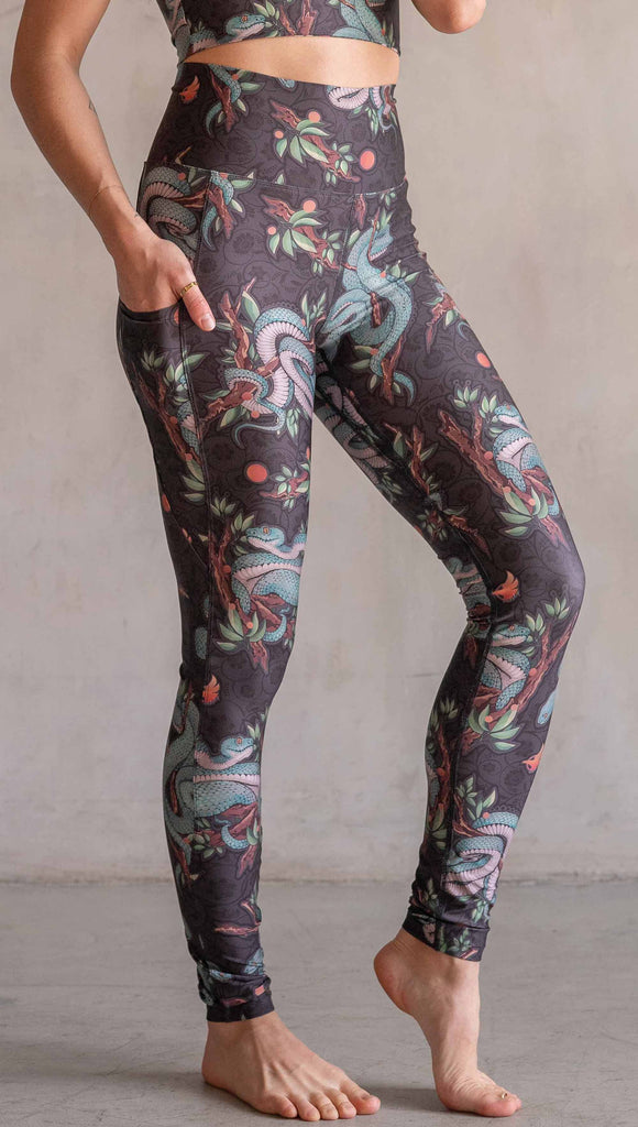 Model wearing WERKSHOP Pit Viper Featherlight Leggings. The artwork on the leggings have clusters of teal blue pit viper snakes intertwined on tree branches over a taupe/brown background. The featherlight leggings have pockets.