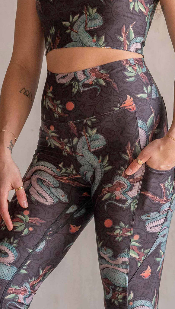 Model wearing WERKSHOP Pit Viper Featherlight Leggings. The artwork on the leggings have clusters of teal blue pit viper snakes intertwined on tree branches over a taupe/brown background. The featherlight leggings have pockets.