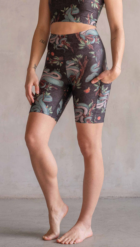 Model wearing WERKSHOP Pit Viper Featherlight Bicycle Length shorts. The artwork on the shorts have clusters of teal blue pit viper snakes intertwined on tree branches over a taupe/brown background. The featherlight shorts have pockets.