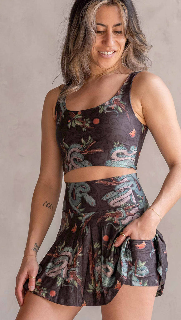 Model wearing WERKSHOP Pit Viper Featherlight Active Skirt. The skirts have built in shorts underneath and artwork on the shorts have clusters of teal blue pit viper snakes intertwined on tree branches over a taupe/brown background. The featherlight skirts have pockets.