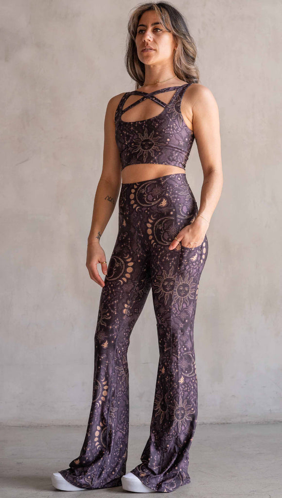 Model wearing WERKSHOP Zodiac Bell Bottoms. They are high waist and feature pockets on both legs. The zodiac themed artwork shows a hand-drawn sun and moon with the moon phases, shooting stars and all 12 zodiac constellations in gold over a dark purple background. Our model is 5’2” and this images shows her wearing the bells with sneakers. The hem of the pants are hitting the floor.