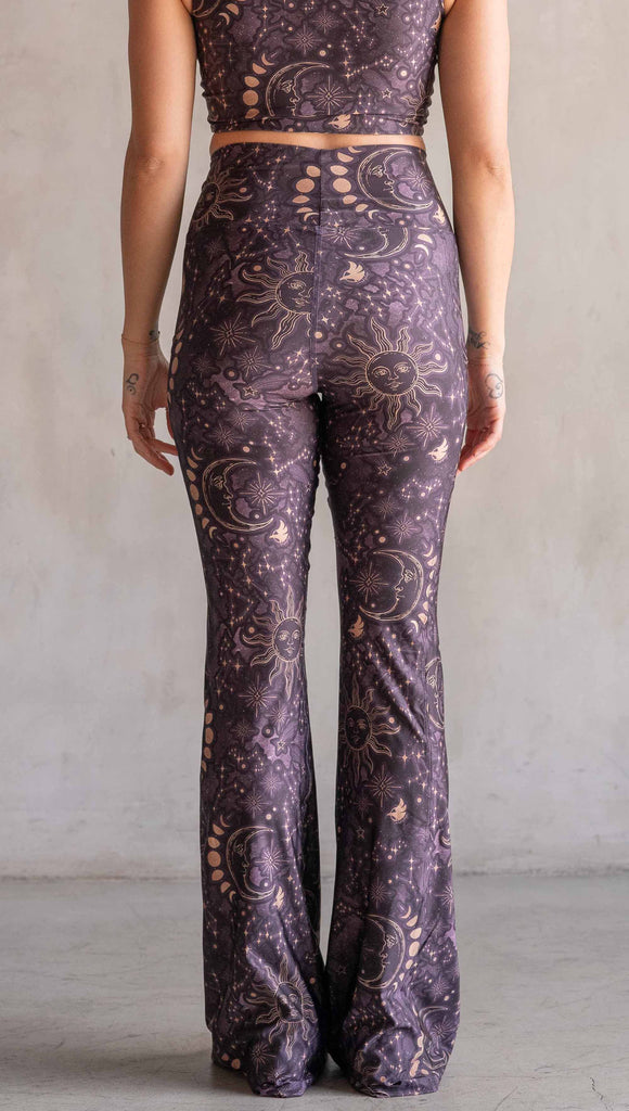 Model wearing WERKSHOP Zodiac Bell Bottoms. They are high waist and feature pockets on both legs. The zodiac themed artwork shows a hand-drawn sun and moon with the moon phases, shooting stars and all 12 zodiac constellations in gold over a dark purple background. Our model is 5’2” and this images shows her wearing the bells with sneakers. The hem of the pants are hitting the floor.