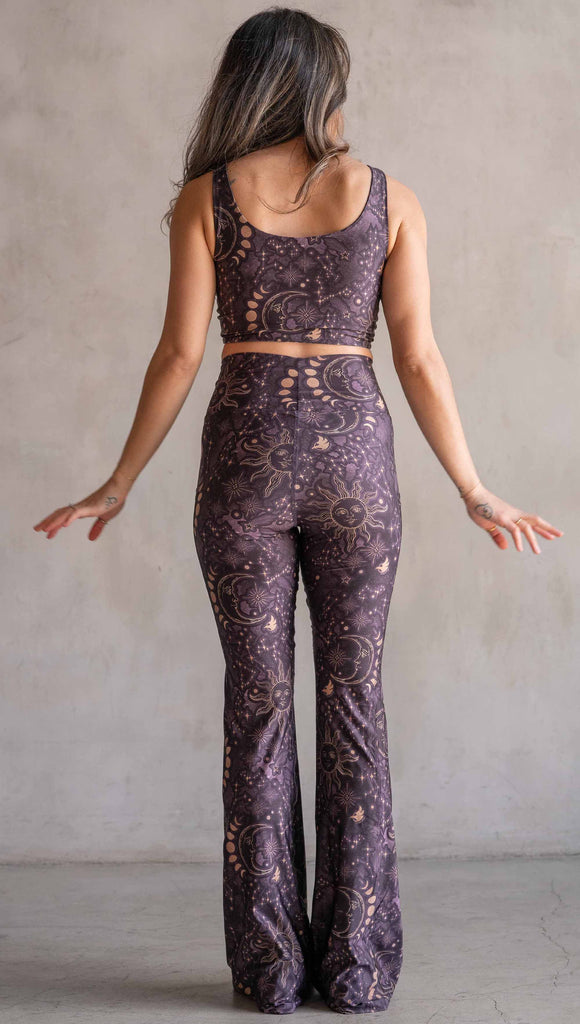 Model wearing WERKSHOP Zodiac Bell Bottoms. They are high waist and feature pockets on both legs. The zodiac themed artwork shows a hand-drawn sun and moon with the moon phases, shooting stars and all 12 zodiac constellations in gold over a dark purple background. Our model is 5’2” and this images shows her wearing the bells with sneakers. The hem of the pants are hitting the floor.