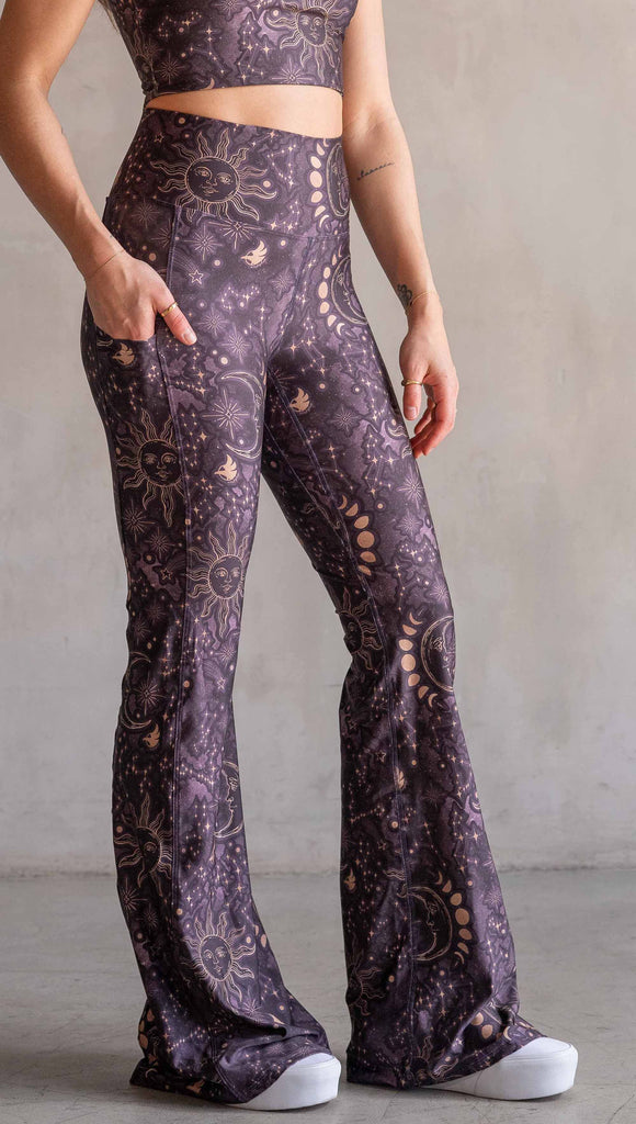 Model wearing WERKSHOP Zodiac Bell Bottoms. They are high waist and feature pockets on both legs. The zodiac themed artwork shows a hand-drawn sun and moon with the moon phases, shooting stars and all 12 zodiac constellations in gold over a dark purple background. Our model is 5’2” and this images shows her wearing the bells with sneakers. The hem of the pants are hitting the floor.