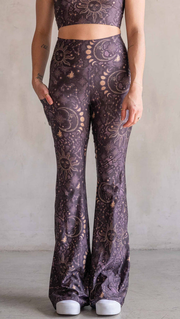 Model wearing WERKSHOP Zodiac Bell Bottoms. They are high waist and feature pockets on both legs. The zodiac themed artwork shows a hand-drawn sun and moon with the moon phases, shooting stars and all 12 zodiac constellations in gold over a dark purple background. Our model is 5’2” and this images shows her wearing the bells with sneakers. The hem of the pants are hitting the floor.