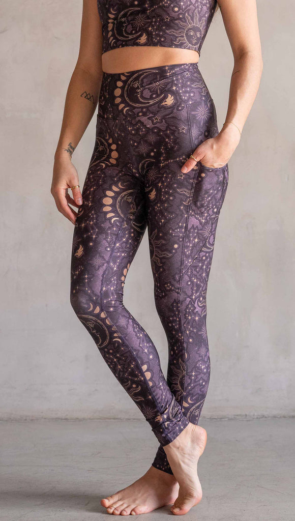 Model wearing WERKSHOP Zodiac Leggings. They are high waist and feature pockets on both legs. The zodiac themed artwork shows a hand-drawn sun and moon with the moon phases, shooting stars and all 12 zodiac constellations in gold over a dark purple background.