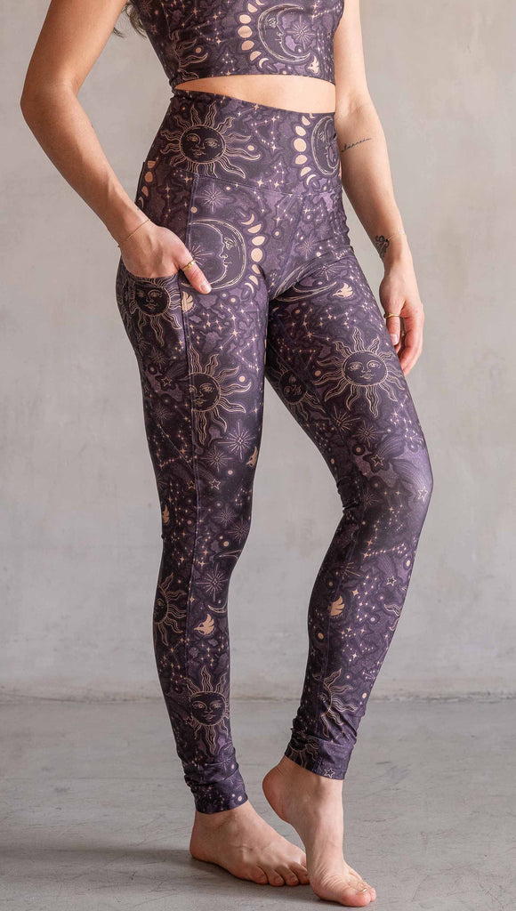 Model wearing WERKSHOP Zodiac Leggings. They are high waist and feature pockets on both legs. The zodiac themed artwork shows a hand-drawn sun and moon with the moon phases, shooting stars and all 12 zodiac constellations in gold over a dark purple background.