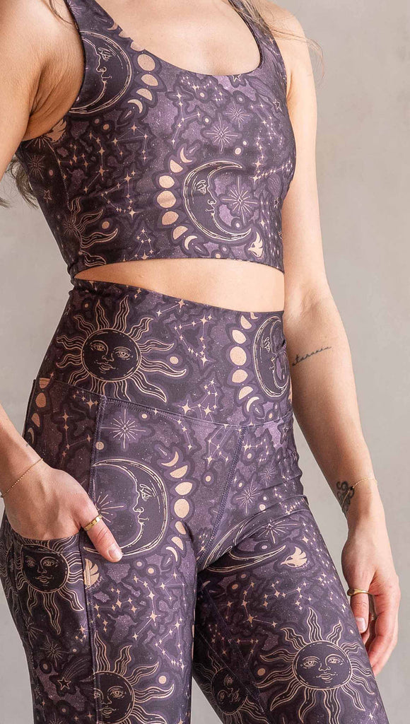 Model wearing WERKSHOP Zodiac Leggings. They are high waist and feature pockets on both legs. The zodiac themed artwork shows a hand-drawn sun and moon with the moon phases, shooting stars and all 12 zodiac constellations in gold over a dark purple background.