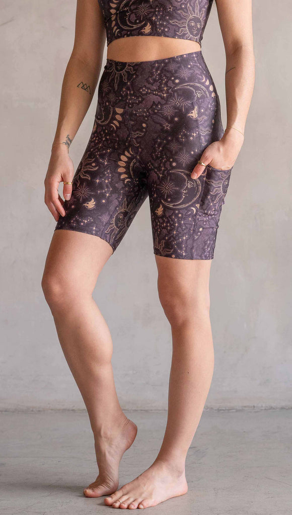 Model wearing WERKSHOP Zodiac Bicycle Length Shorts. They are high waist and feature pockets on both legs. The zodiac themed artwork shows a hand-drawn sun and moon with the moon phases, shooting stars and all 12 zodiac constellations in gold over a dark purple background. The length of the shorts hit in the mid thigh, above the knee.