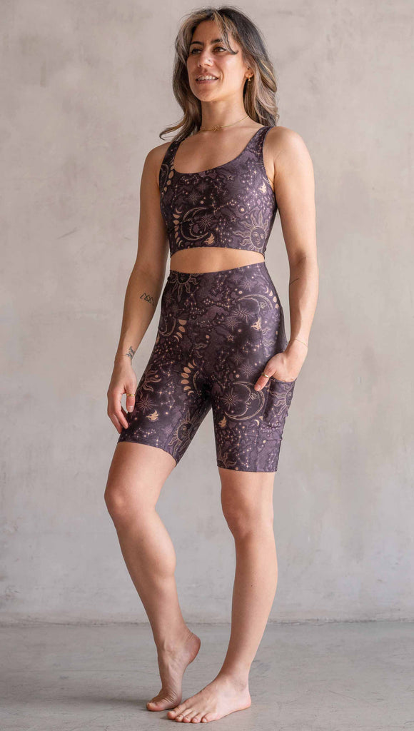 Model wearing WERKSHOP Zodiac Bicycle Length Shorts. They are high waist and feature pockets on both legs. The zodiac themed artwork shows a hand-drawn sun and moon with the moon phases, shooting stars and all 12 zodiac constellations in gold over a dark purple background. The length of the shorts hit in the mid thigh, above the knee.