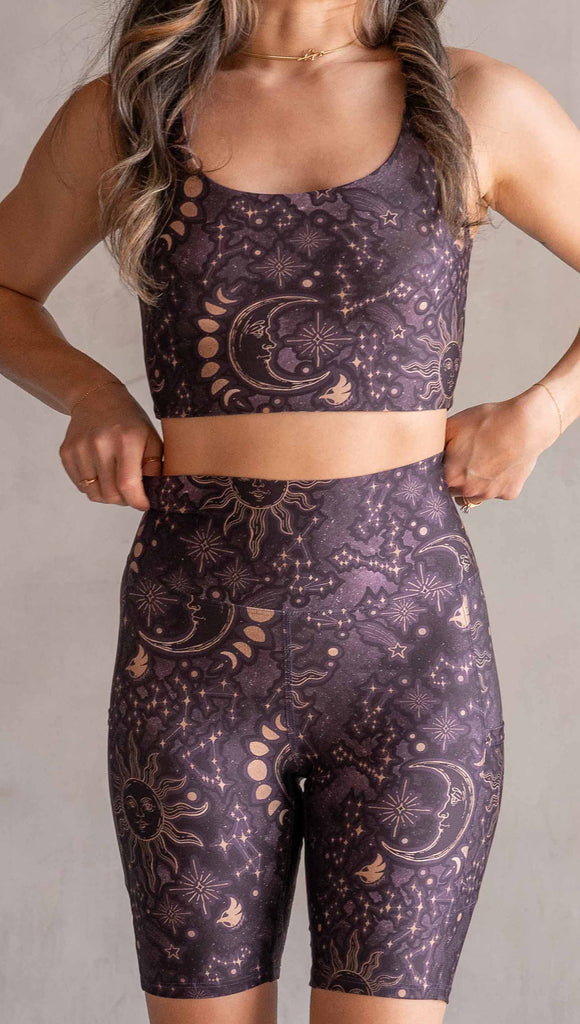 Model wearing WERKSHOP Zodiac Bicycle Length Shorts. They are high waist and feature pockets on both legs. The zodiac themed artwork shows a hand-drawn sun and moon with the moon phases, shooting stars and all 12 zodiac constellations in gold over a dark purple background. The length of the shorts hit in the mid thigh, above the knee.