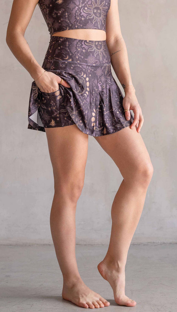 Model wearing WERKSHOP Zodiac Tennis Skirt with built-in shorts. They are high waist and feature pockets on both legs under the flirty skirt. The zodiac themed artwork shows a hand-drawn sun and moon with the moon phases, shooting stars and all 12 zodiac constellations in gold over a dark purple background. The skirt length hits right around the models fingertips.