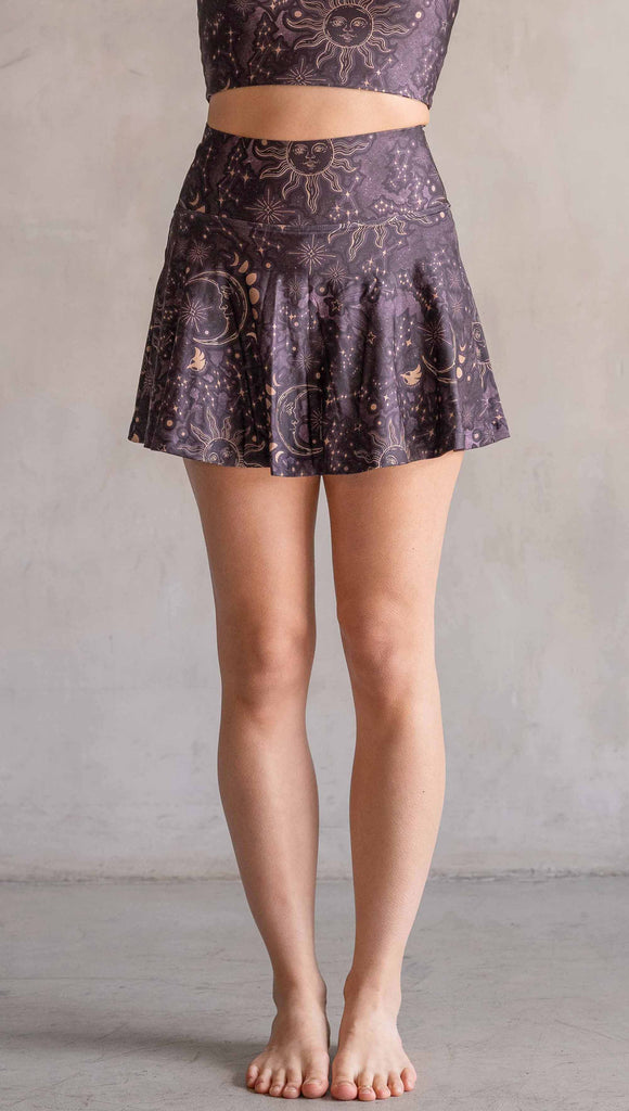 Model wearing WERKSHOP Zodiac Tennis Skirt with built-in shorts. They are high waist and feature pockets on both legs under the flirty skirt. The zodiac themed artwork shows a hand-drawn sun and moon with the moon phases, shooting stars and all 12 zodiac constellations in gold over a dark purple background. The skirt length hits right around the models fingertips.