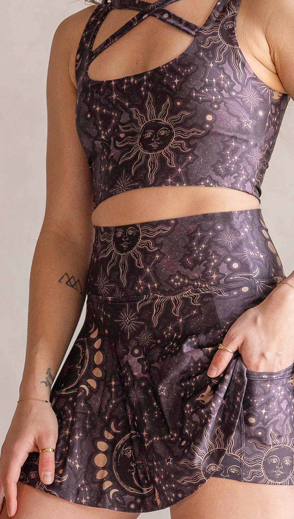 Model wearing WERKSHOP Zodiac Tennis Skirt with built-in shorts. They are high waist and feature pockets on both legs under the flirty skirt. The zodiac themed artwork shows a hand-drawn sun and moon with the moon phases, shooting stars and all 12 zodiac constellations in gold over a dark purple background. The skirt length hits right around the models fingertips.