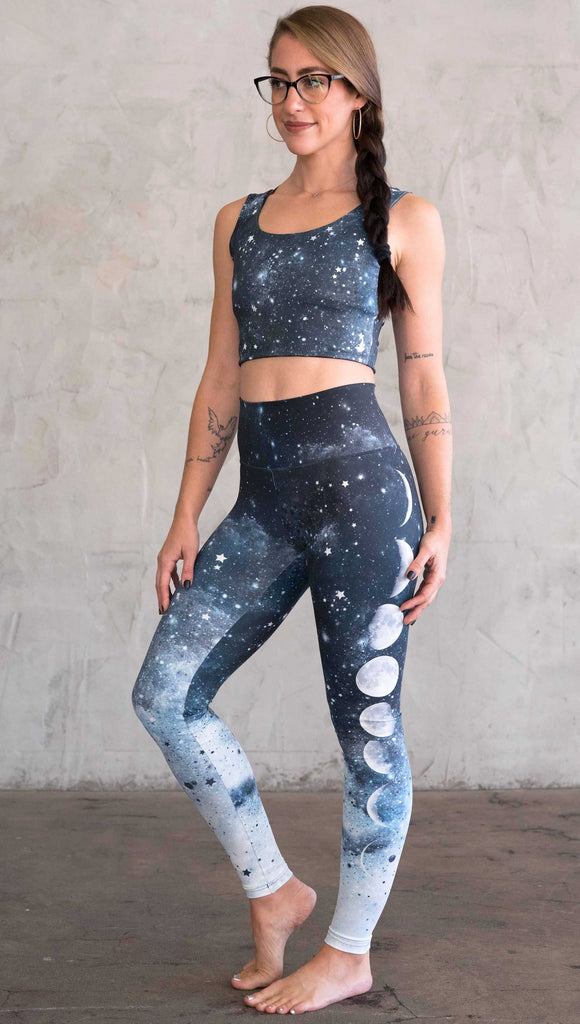 full body view of a girl wearing our moon phases athleisure leggings - with the phases of the moon printed on the wearer's left leg on a starry night sky with all zodiac constellations in the background