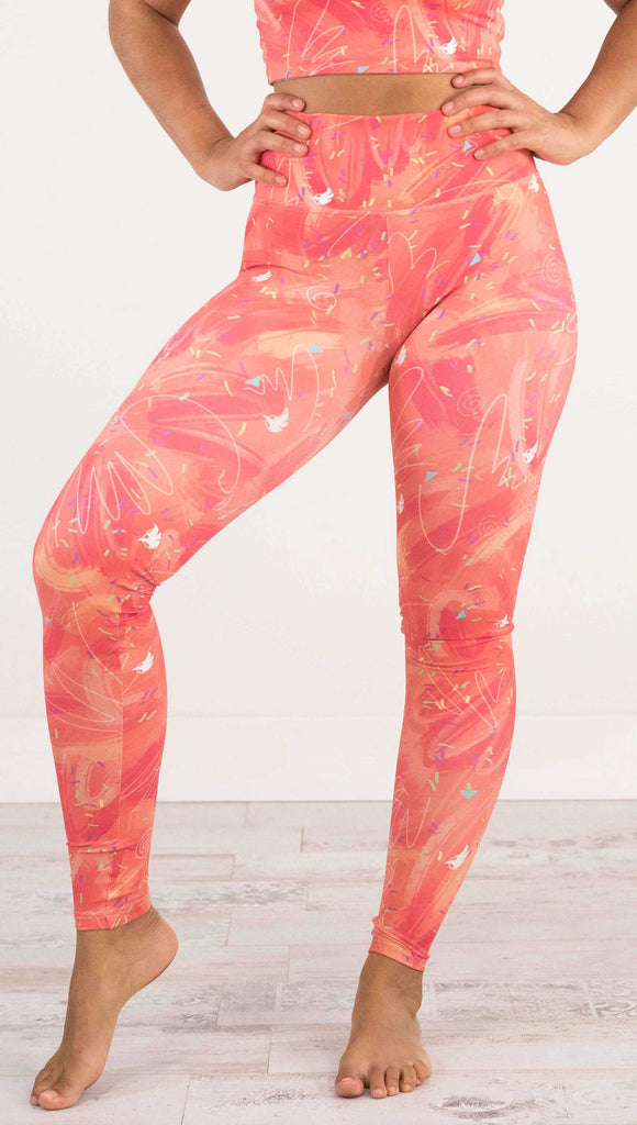 Waist down front view of model wearing WERKSHOP Salmon Scribble Leggings with coral  brushstrokes over a bright salmon background. Also has little confetti and eagle logos scattered throughout.