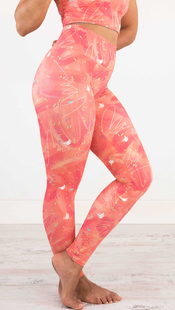 Waist down side view of model wearing WERKSHOP Salmon Scribble Leggings with coral  brushstrokes over a bright salmon background. Also has little confetti and eagle logos scattered throughout.