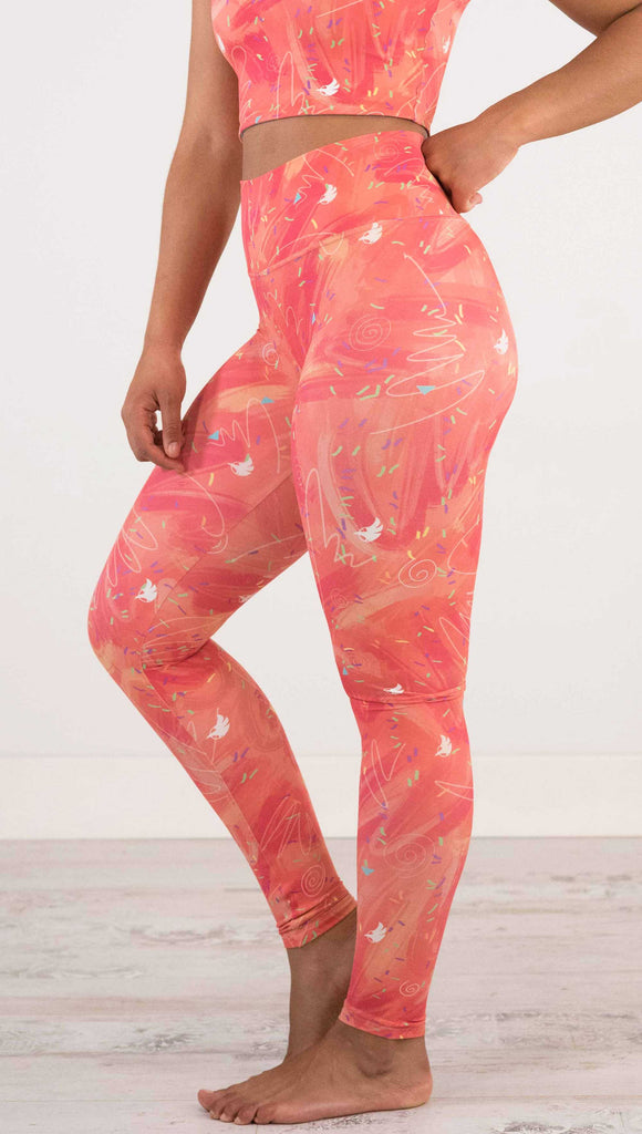 Waist down side view of model wearing WERKSHOP Salmon Scribble Leggings with coral  brushstrokes over a bright salmon background. Also has little confetti and eagle logos scattered throughout.