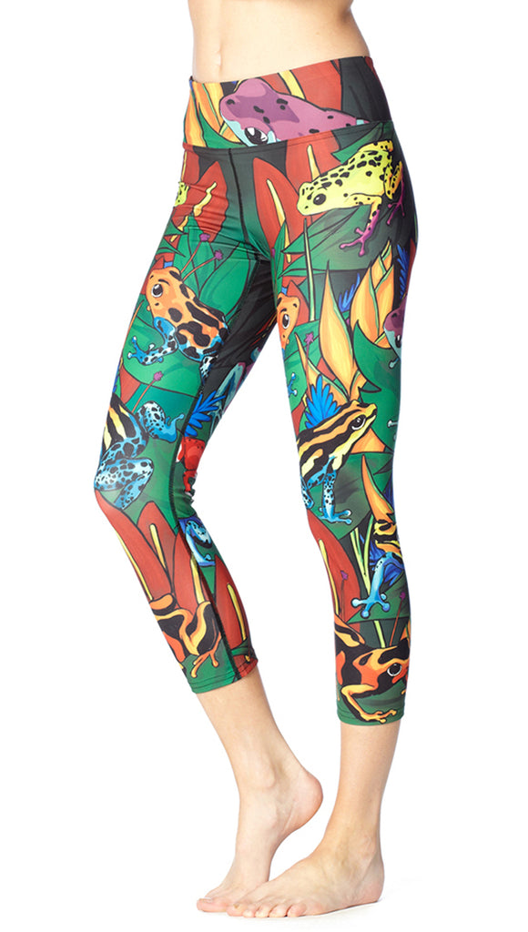 Model wearing leggings printed with poison dark frog artwork. The leggings are mostly green with pops of bright red, yellow and blow. The frogs are camouflaged in foliage and flowers.