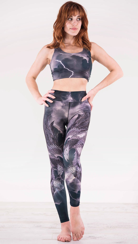 Front view of model wearing dark gray triathlon leggings with a large purple bird across each leg with lightning in the background