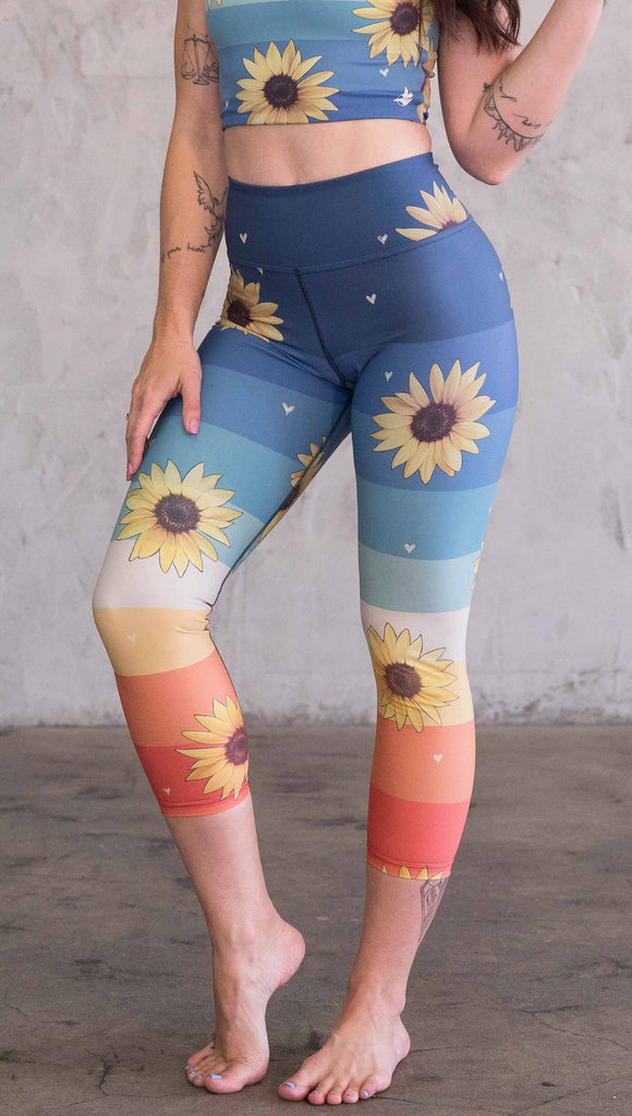 Waist down front view of model wearing WERKSHOP Sunflower triathlon capri leggings. The leggings have wide horizontal stripes with dark blue at the waistband, to aqua and pale green at the mid thigh leading to cream at the knee and orange and red tones to the ankle. There are large photo-real sunflowers and tiny hand sketched off-white hearts sprinkled throughout.