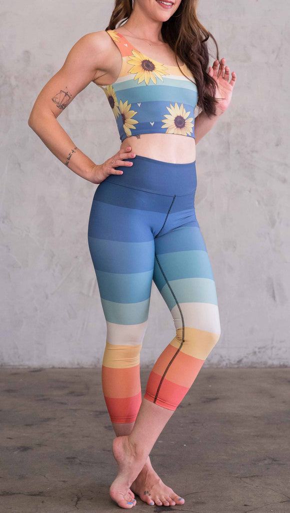 Full body front view of model wearing WERKSHOP Vintage Rainbow Triathlon Capri leggings. The leggings have wide horizontal stripes with dark blue at the waistband, to auqua and pale green at the mid thigh leading to cream at the knee and orange and red tones to the ankle.